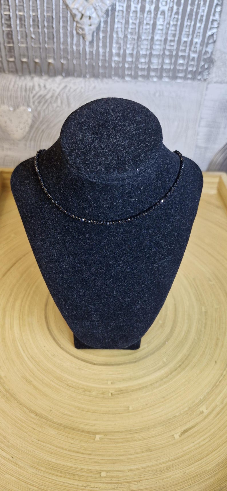 May include: A delicate black beaded necklace displayed on a black velvet jewelry bust. The necklace is composed of small, evenly spaced beads, creating a simple yet elegant design. The bust sits on a light brown wooden surface.