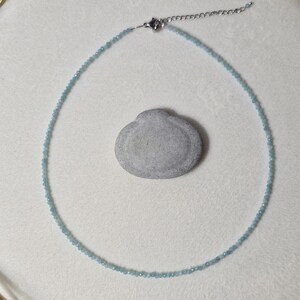 May include: A delicate necklace made of small, light blue beads. The necklace is on a white surface with a small, gray rock.