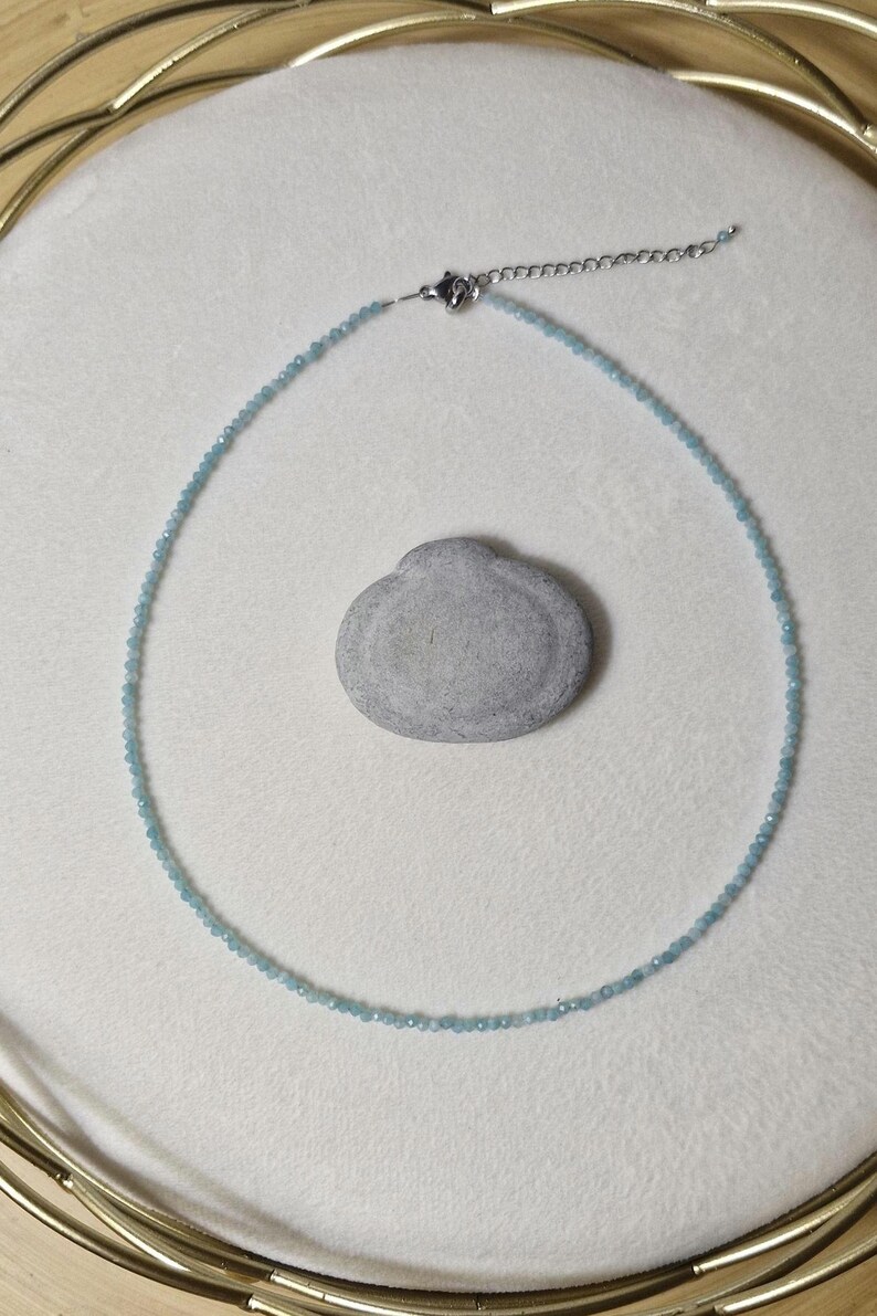 May include: A delicate necklace made of small, light blue beads. The necklace is on a white surface with a small, gray rock.