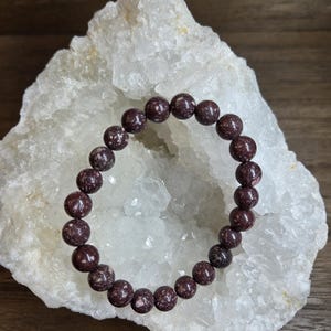 Peut inclure: Un bracelet composé de perles sphériques de couleur marron foncé avec des motifs tachetés blancs. Les perles sont enfilées en une boucle continue, présentant une texture lisse et polie sur l'ensemble de ce bijou circulaire.
