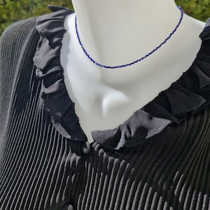 May include: A delicate, dark blue beaded necklace displayed on a mannequin. The necklace is a simple strand of small, evenly spaced beads. The mannequin is wearing a black, pleated blouse with a ruffled collar.