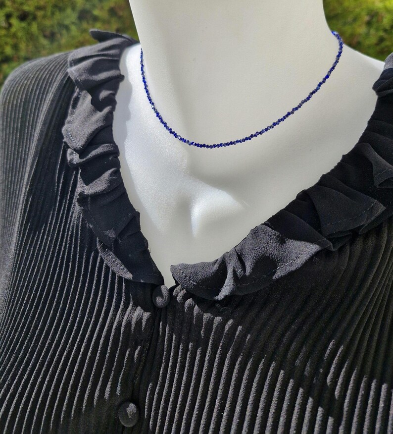 May include: A delicate, dark blue beaded necklace displayed on a mannequin. The necklace is a simple strand of small, evenly spaced beads. The mannequin is wearing a black, pleated blouse with a ruffled collar.