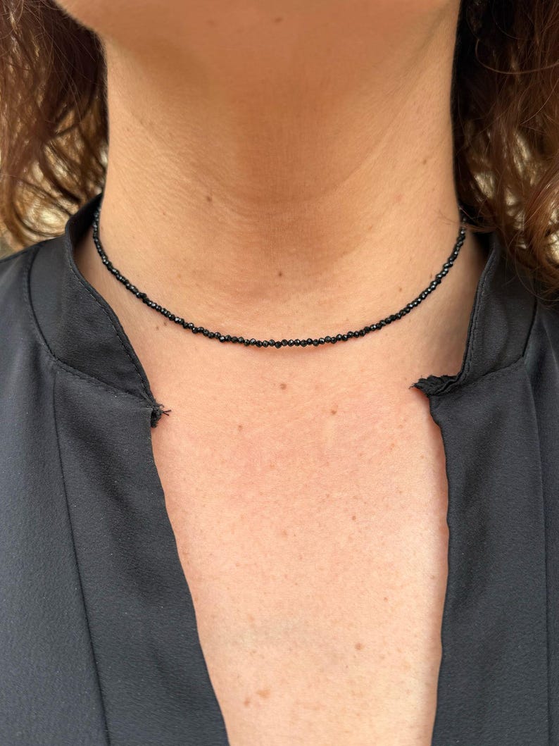 May include: A fine black beaded choker necklace. The necklace is made of small, dark beads, forming a close-fitting collar. The necklace is worn against a dark-coloured top.