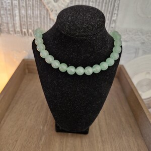 May include: A light green beaded necklace on a black velvet display stand. The necklace is made of round beads and is approximately 18 inches long.