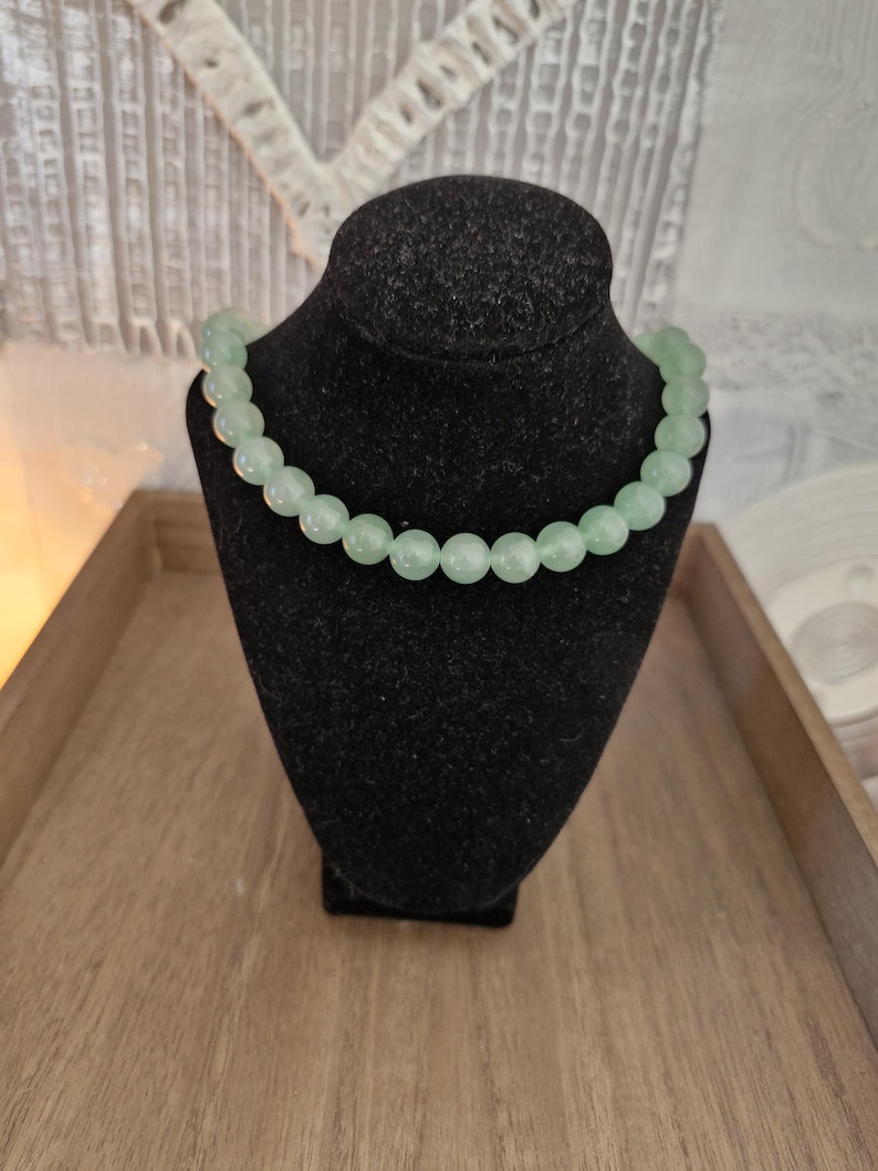 May include: A light green beaded necklace on a black velvet display stand. The necklace is made of round beads and is approximately 18 inches long.