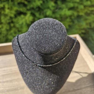 May include: A fine black beaded necklace displayed on a black velvet jewellery bust. The necklace is a thin strand of small, dark beads. The bust is set on a light-coloured wooden surface, with a blurred green background.