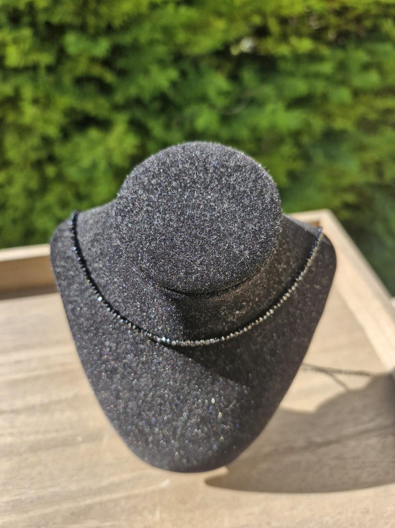 May include: A fine black beaded necklace displayed on a black velvet jewellery bust. The necklace is a thin strand of small, dark beads. The bust is set on a light-coloured wooden surface, with a blurred green background.