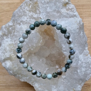 Könnte beinhalten: Ein grünes und weißes Agat-Armband auf einer weißen Kristall-Geode. Das Armband besteht aus runden Perlen mit einem natürlichen Steinmuster.