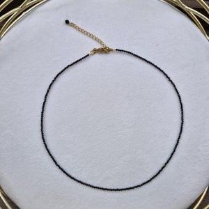 May include: A fine black beaded necklace with a gold-coloured clasp and chain. The necklace is displayed on a white surface, with a gold-coloured decorative frame in the background. The necklace is a choker style.