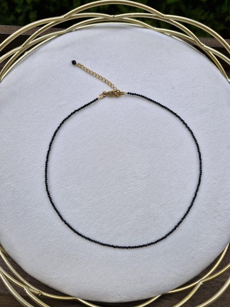 May include: A fine black beaded necklace with a gold-coloured clasp and chain. The necklace is displayed on a white surface, with a gold-coloured decorative frame in the background. The necklace is a choker style.