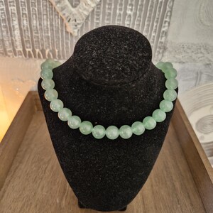 May include: A light green beaded necklace made of round, smooth stones. The necklace is displayed on a black velvet mannequin bust.