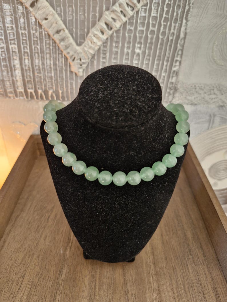 May include: A light green beaded necklace made of round, smooth stones. The necklace is displayed on a black velvet mannequin bust.