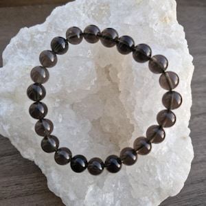 May include: A black smoky quartz beaded bracelet with a dark grey colour. The bracelet is made of round beads and is lying on a white rock.