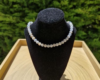 May include: A beaded necklace with light-colored, round beads is displayed on a black velvet bust. The necklace is draped around the bust, showcasing its design. The background features green foliage and a wooden tray.