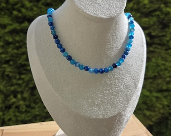 Puede incluir: Un collar de cuentas que presenta una serie de pequeñas piedras redondas y pulidas en varios tonos de azul, que van desde el azul cielo claro hasta el azul marino profundo, dispuestas en un patrón repetitivo a lo largo del hilo circular.