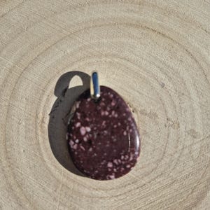 Peut inclure: Un pendentif marron avec des mouchetures blanches et une bélière argentée. La pierre polie en forme de goutte d'eau est présentée sur une surface en bois brun clair.