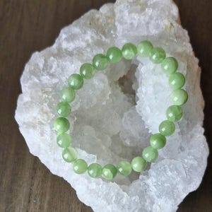 Peut inclure: Un bracelet en jade vert avec des perles rondes, reposant sur un amas de cristaux blancs.