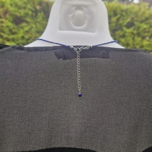 May include: A dark blue beaded necklace with a silver chain and clasp. The necklace is displayed on a white mannequin head, against a backdrop of green foliage. The necklace has a small blue bead at the end of the chain.