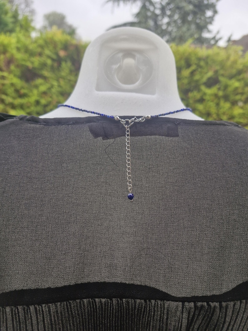 May include: A dark blue beaded necklace with a silver chain and clasp. The necklace is displayed on a white mannequin head, against a backdrop of green foliage. The necklace has a small blue bead at the end of the chain.