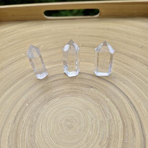 Può includere: Tre punte di cristallo di quarzo trasparente su un vassoio di legno. Le punte sono tutte della stessa dimensione e forma, e sono disposte in fila.