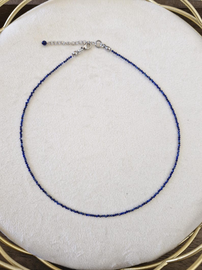 May include: Delicate necklace featuring small, deep blue beads strung together. The necklace has a silver-toned clasp and adjustable chain. The jewelry is displayed on a white surface.