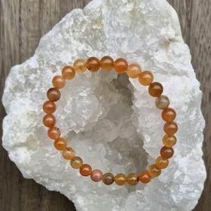 Puede incluir: Un brazalete hecho de cuentas de piedras preciosas de color naranja y marrón. El brazalete está sobre un cúmulo de cristales blancos.