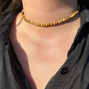 Op de afbeelding: Een kralen choker ketting met kleine, ronde, aardetinten kralen. De ketting wordt gedragen tegen een donker overhemd, waardoor een contrast ontstaat dat de natuurlijke kleuren van de kralen benadrukt. De kralen zijn ongeveer 0,6 cm in diameter.