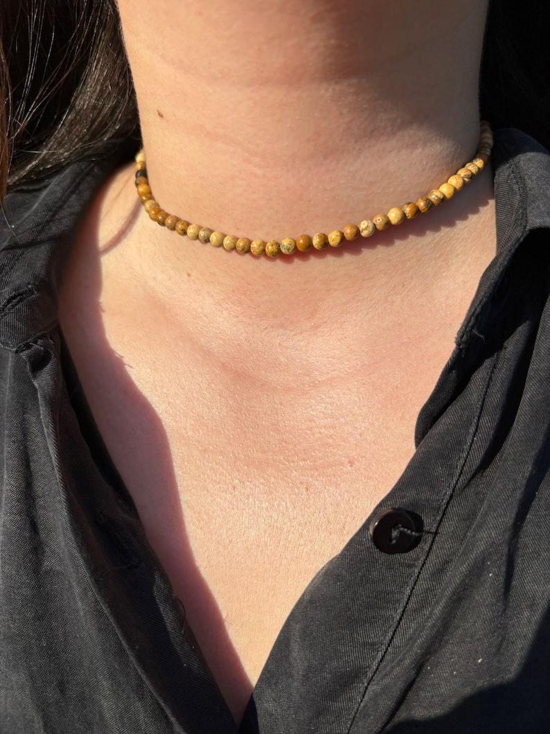 Op de afbeelding: Een kralen choker ketting met kleine, ronde, aardetinten kralen. De ketting wordt gedragen tegen een donker overhemd, waardoor een contrast ontstaat dat de natuurlijke kleuren van de kralen benadrukt. De kralen zijn ongeveer 0,6 cm in diameter.