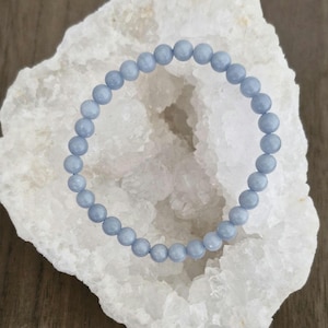 Könnte beinhalten: Ein hellblaues Edelstein-Armband mit runden Perlen. Das Armband wird auf einer weißen Kristall-Geode präsentiert.