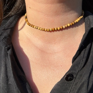 Op de afbeelding: Een choker ketting met kleine, ronde kralen in lichtbruine en beige tinten. De kralen zijn aan elkaar geregen om een delicate ketting te vormen. De ketting is een eenvoudig, elegant accessoire, gedragen om de nek.