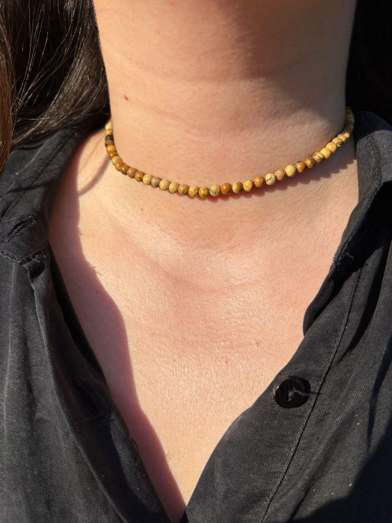 Op de afbeelding: Een choker ketting met kleine, ronde kralen in lichtbruine en beige tinten. De kralen zijn aan elkaar geregen om een delicate ketting te vormen. De ketting is een eenvoudig, elegant accessoire, gedragen om de nek.