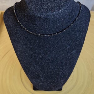 May include: A black beaded necklace displayed on a black velvet bust. The necklace is made of small, dark beads, giving it a delicate and elegant look. The bust is set against a light wooden background.