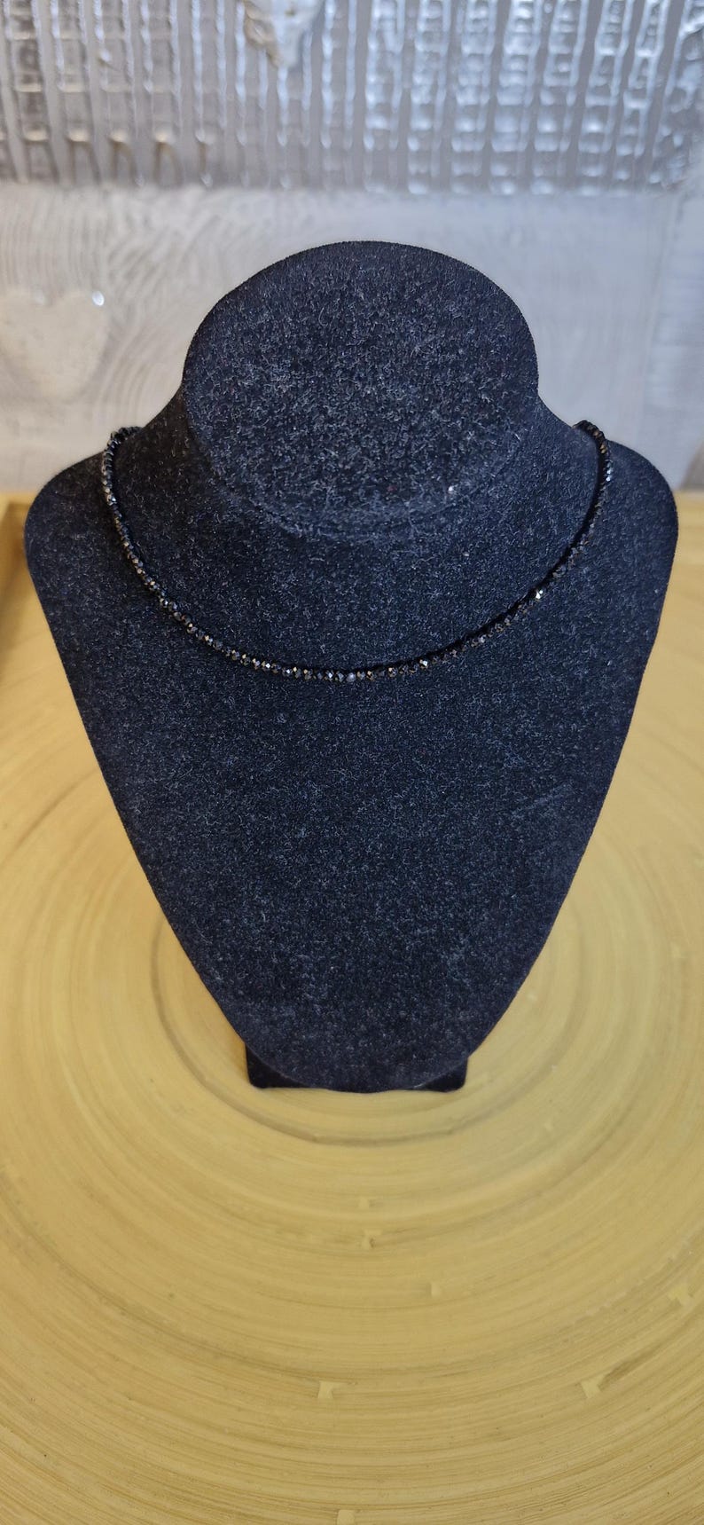May include: A black beaded necklace displayed on a black velvet bust. The necklace is made of small, dark beads, giving it a delicate and elegant look. The bust is set against a light wooden background.