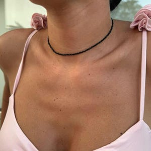 May include: A fine black beaded choker necklace. The necklace is worn around the neck. The beads are small and evenly spaced. The person is wearing a light pink spaghetti strap top.