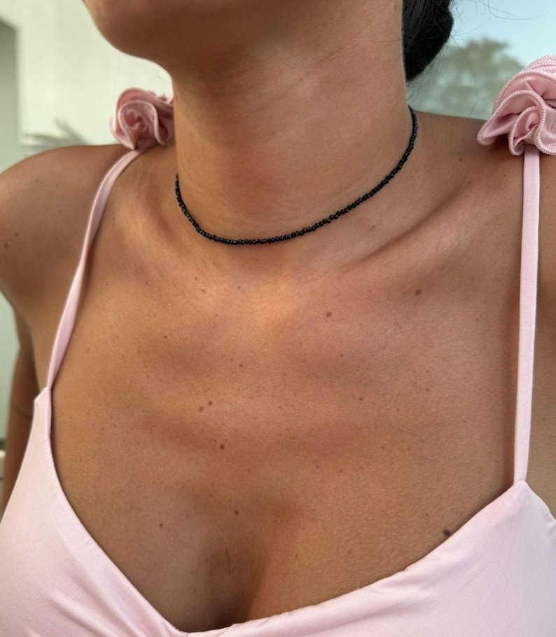 May include: A fine black beaded choker necklace. The necklace is worn around the neck. The beads are small and evenly spaced. The person is wearing a light pink spaghetti strap top.