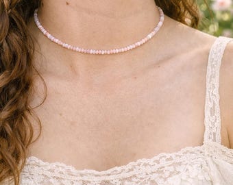 Puede incluir: Un delicado collar de cuentas rosas. El collar está compuesto por pequeñas cuentas redondas, creando un aspecto sutil y elegante. El collar se lleva alrededor del cuello. El fondo está borroso, lo que sugiere un entorno al aire libre.