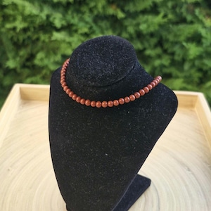 May include: A necklace with small, round, reddish-brown beads is displayed on a black velvet bust. The necklace is draped around the bust, showcasing its design. The background features green foliage and a wooden tray.