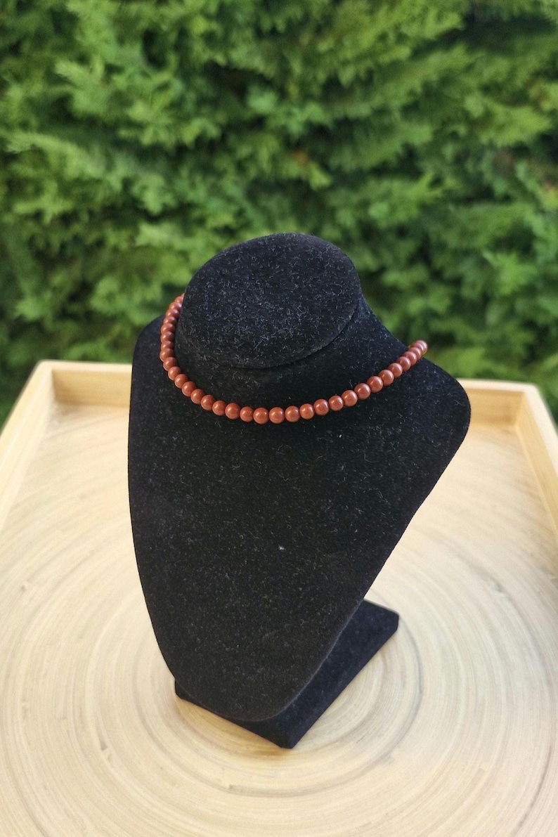May include: A necklace with small, round, reddish-brown beads is displayed on a black velvet bust. The necklace is draped around the bust, showcasing its design. The background features green foliage and a wooden tray.