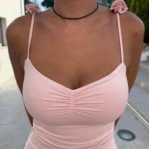 May include: A pale pink spaghetti strap dress with a gathered bodice. The dress has small rose details on the straps and is worn with a black beaded choker necklace. A small heart tattoo is visible on the shoulder.