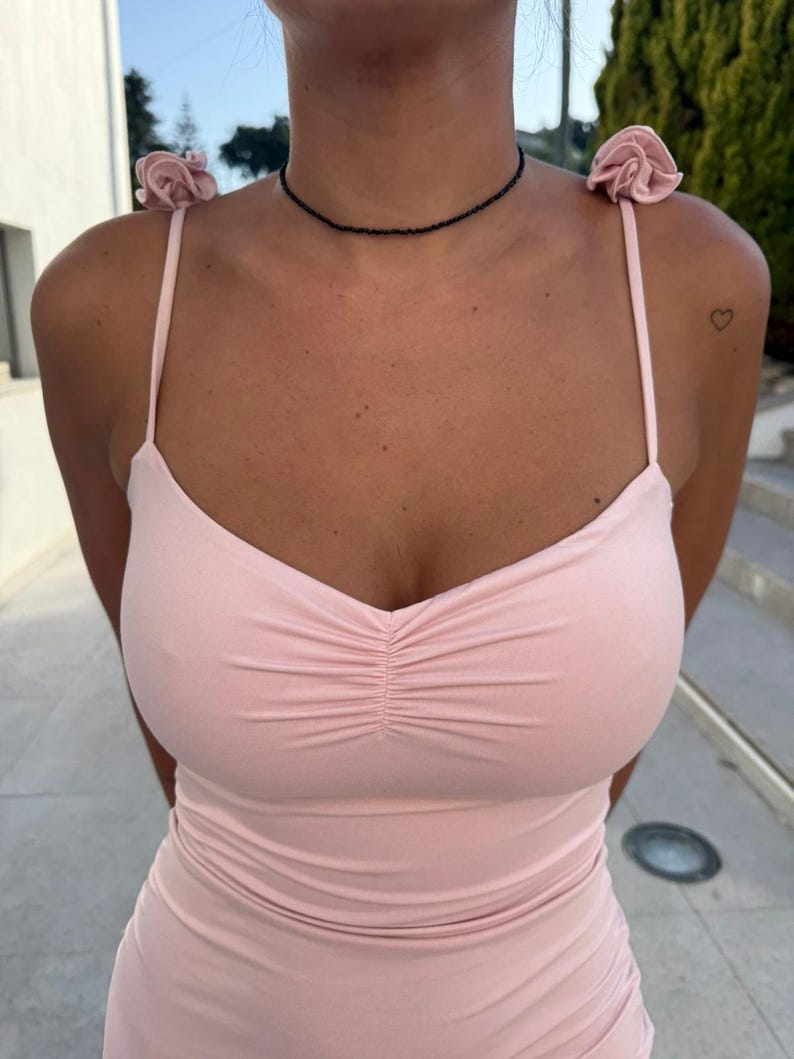 May include: A pale pink spaghetti strap dress with a gathered bodice. The dress has small rose details on the straps and is worn with a black beaded choker necklace. A small heart tattoo is visible on the shoulder.