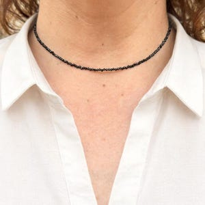 May include: A black beaded choker necklace. The necklace is made of small, dark beads, creating a simple yet elegant accessory. The necklace is worn against a white collared shirt.