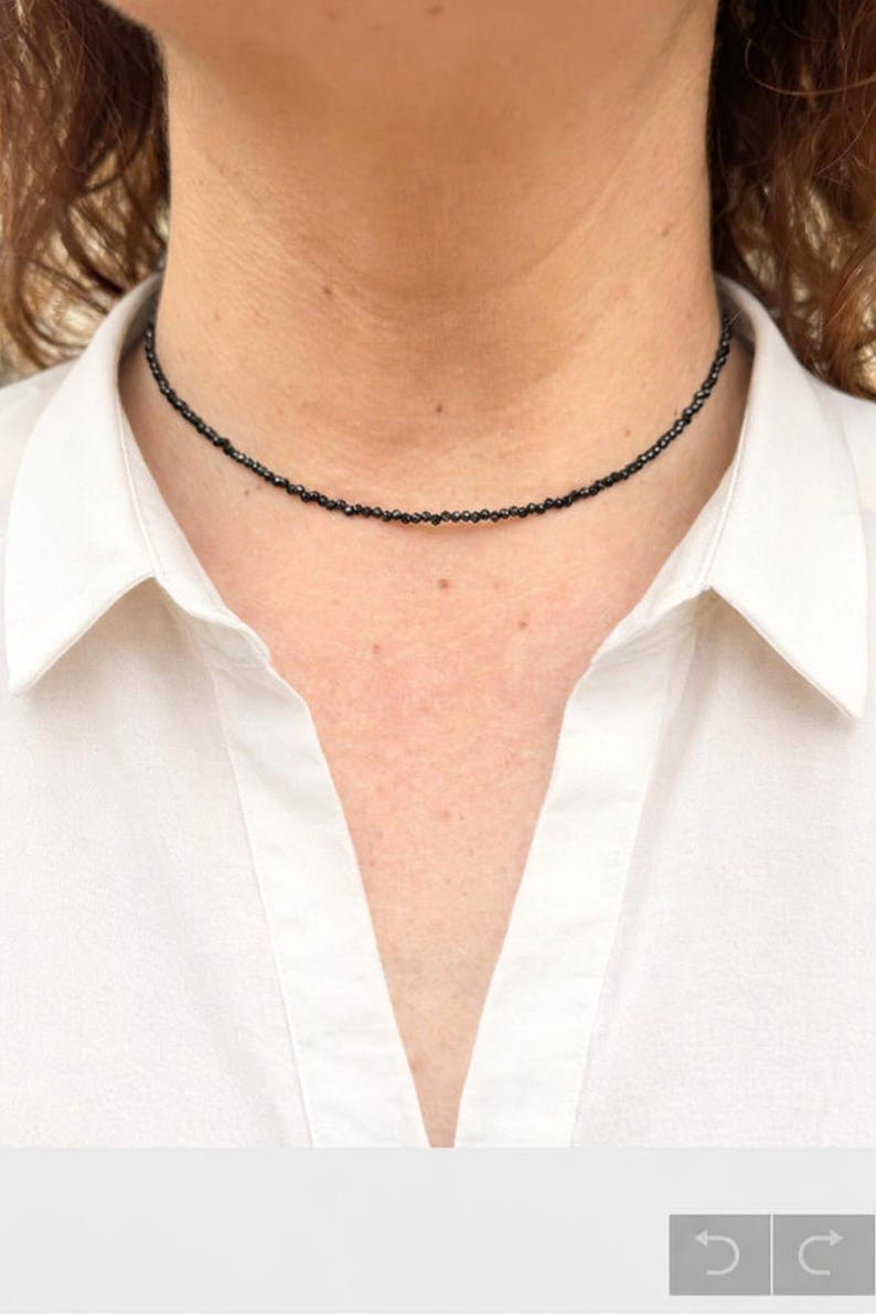 May include: A black beaded choker necklace. The necklace is made of small, dark beads, creating a simple yet elegant accessory. The necklace is worn against a white collared shirt.