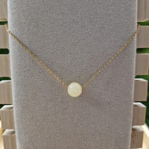 Op de afbeelding: Een delicate goudkleurige ketting met een enkele, ronde, cr&egrave;mekleurige kralen hanger. De ketting wordt gepresenteerd op een grijze fluwelen ondergrond, die de eenvoudige elegantie benadrukt. De ketting is dun en de kraal is gecentreerd.