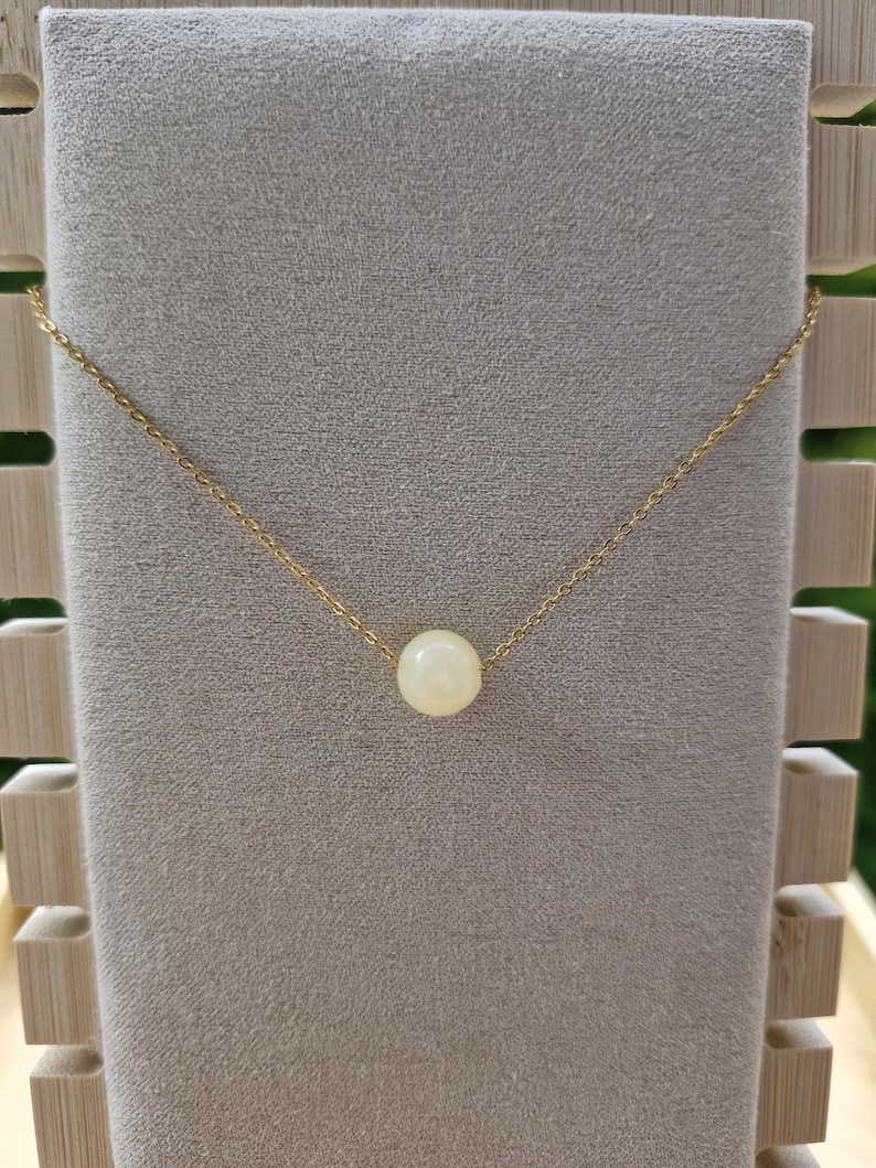 Op de afbeelding: Een delicate goudkleurige ketting met een enkele, ronde, cr&egrave;mekleurige kralen hanger. De ketting wordt gepresenteerd op een grijze fluwelen ondergrond, die de eenvoudige elegantie benadrukt. De ketting is dun en de kraal is gecentreerd.