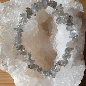 Peut inclure: Un bracelet composé de petites pierres de labradorite grises de forme irrégulière. Les pierres sont enfilées sur un élastique. Le bracelet est présenté sur une surface cristalline blanche.