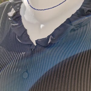 May include: A delicate, dark blue beaded necklace displayed on a mannequin. The necklace is a choker style, with small, evenly spaced beads. The mannequin is wearing a dark blue, pleated blouse with a ruffled collar and button details.