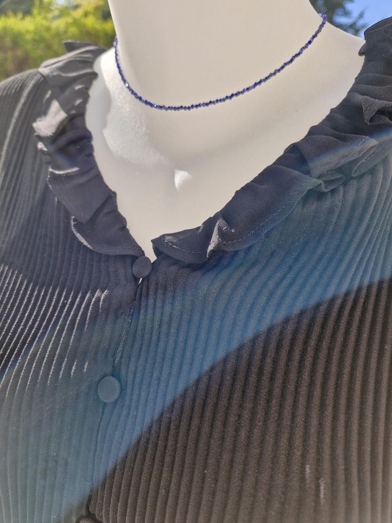 May include: A delicate, dark blue beaded necklace displayed on a mannequin. The necklace is a choker style, with small, evenly spaced beads. The mannequin is wearing a dark blue, pleated blouse with a ruffled collar and button details.