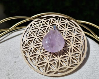 May include: A teardrop-shaped, light purple amethyst pendant hangs above a wooden, circular coaster with a flower of life design. The coaster is light brown, and the pendant is suspended by a silver-colored bail.