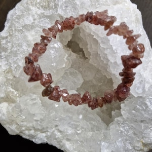 May include: A bracelet made of reddish-brown gemstones, each stone is small and irregularly shaped. The bracelet is set against a white, crystalline geode. The contrast highlights the bracelet's color and texture. The stones appear polished.
