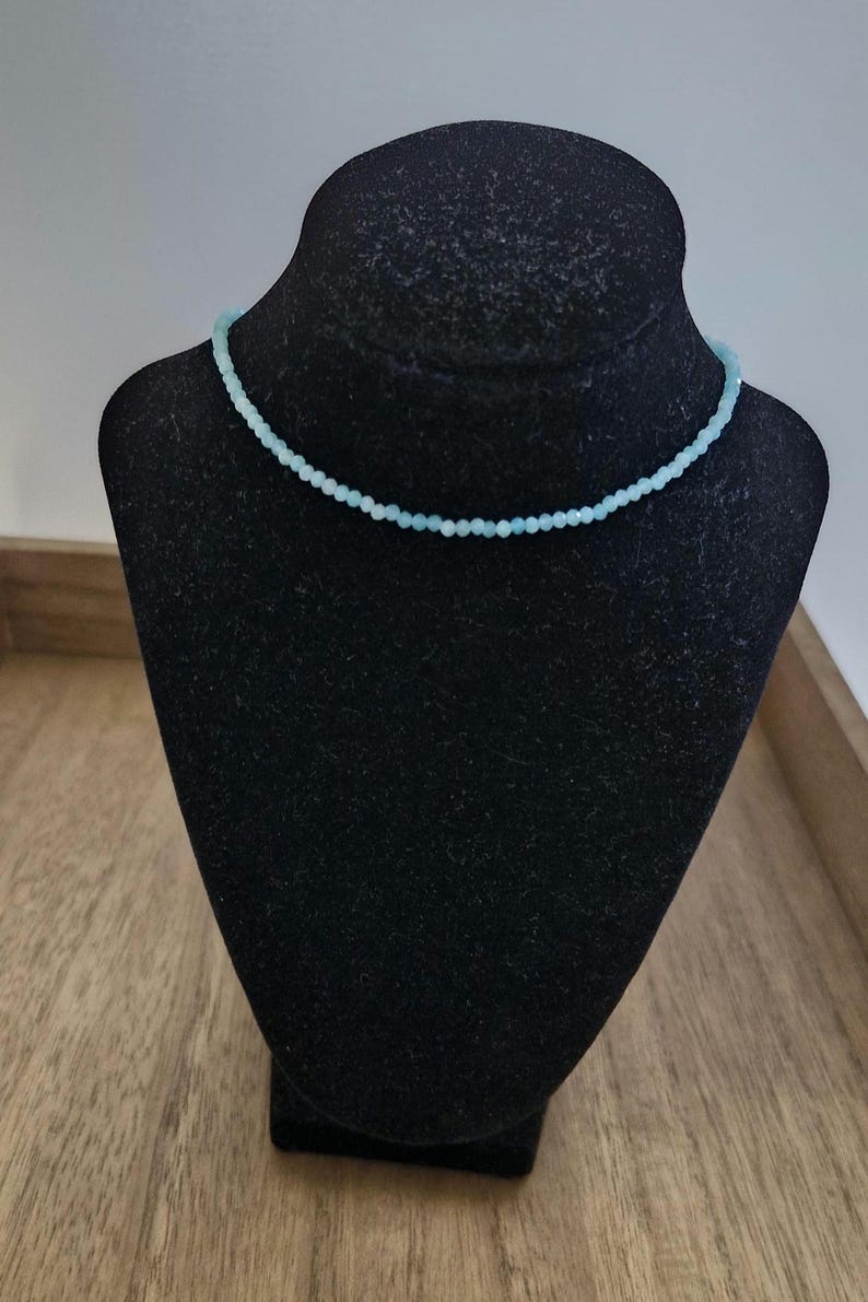 May include: A delicate light blue beaded necklace on a black velvet display stand.